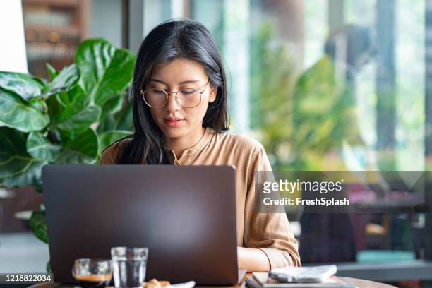 online business: young asian businesswoman working remotely on her laptop computer in a coffee shop - writing email stock pictures, royalty-free photos & images
