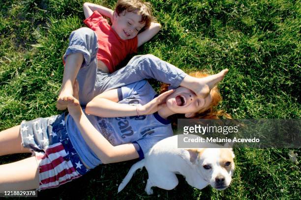 young redheaded girl short hair brown hair laying on her back on grass wrestling younger brother outside on sunny day feet up brother shoving his foot in sisters foot both smiling
