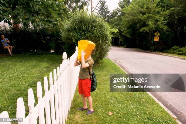 young boy with brown curly hair brown eyes holding yellow cheese hat to his face standing next to fence outside far away from camera - brown hat imagens e fotografias de stock