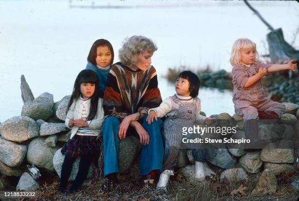 Portrait of American actress Mia Farrow as she poses outdoors, seated on a low wall, with her children, Martha's Vineyard, Massachusetts, 1979....