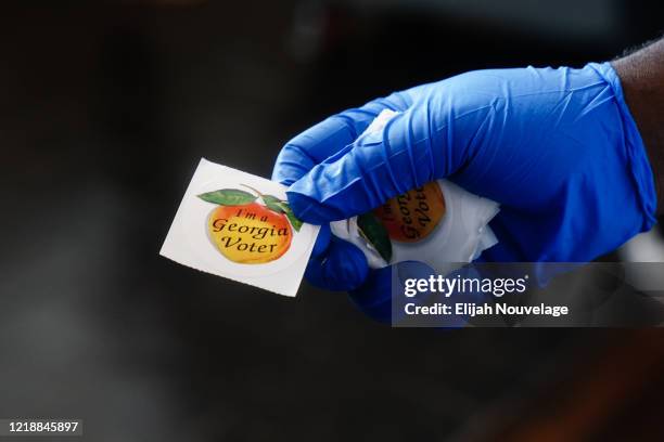 Polling place worker holds an "I'm a Georgia Voter" sticker to hand to a voter on June 9, 2020 in Atlanta, Georgia. Georgia, West Virginia, South...