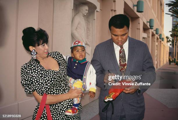 Yolanda Ali holding her adopted son, Asaad Ali, as husband, American heavyweight boxer Muhammad Ali autographs a boxing glove, circa 1986.