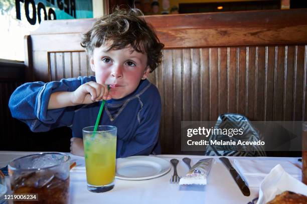 little boy in a restaurant sitting in a booth, drinking from a large glass of orange juice with a green straw, which is on the table. he has brown curly hair and brown eyes and his face is sunburned. - straw stock pictures, royalty-free photos & images