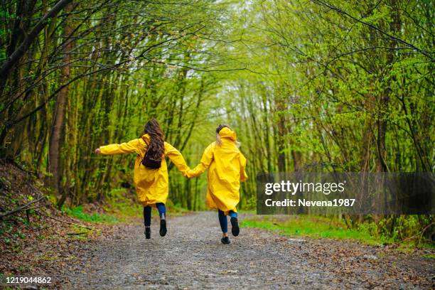 two female friends in raincoats having fun while running on rain in foggy forest - abril imagens e fotografias de stock