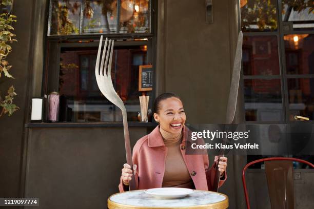 happy woman holding large fork and table knife while sitting at sidewalk cafe - cuisine humour stock-fotos und bilder