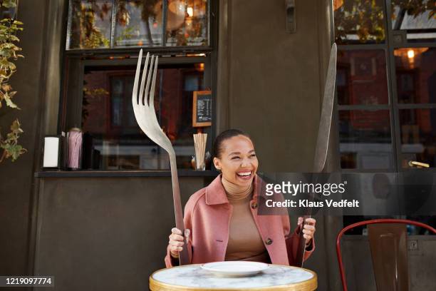 happy woman holding large fork and table knife while sitting at sidewalk cafe - tafelmes stockfoto's en -beelden