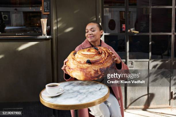 smiling woman holding large raisin roll while sitting at sidewalk cafe - larger than life stock-fotos und bilder