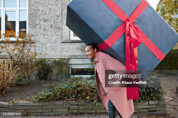 cheerful woman carrying large gift box on footpath - funny christmas gift stockfoto's en -beelden