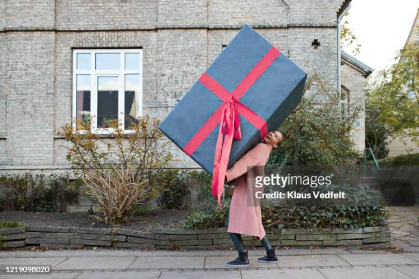 happy woman carrying large gift box on footpath - cadeau noel stockfoto's en -beelden