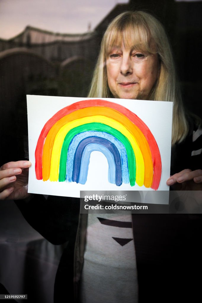 Senior woman holding rainbow drawing in window in support of national health service during Covid-19