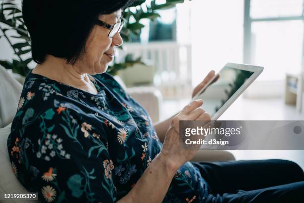 senior asian woman using digital tablet while relaxing on the sofa in the living room at home - asian watching movie stock-fotos und bilder
