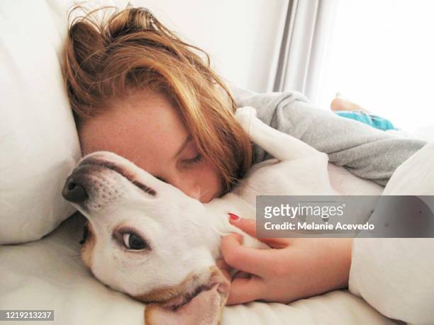 Little redheaded girl laying in bed with her white dog. She is hugging the dog and her eyes are closed. The dog is looking back at the camera and she looks like she is smiling at the camera.
