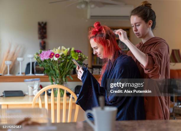 older sister cutting her younger sister's hair at home in the living room, while she is video chatting with her friends and streaming the process of the haircut online. - alex potemkin coronavirus stock pictures, royalty-free photos & images