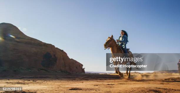 a teenaged native american girl (navajo) rides her horse through the monument valley desert in arizona/utah next to a large rock formation on a clear, bright day - horseback riding arizona stock pictures, royalty-free photos & images