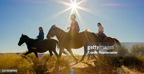 several young native american (navajo) children ride horses through the monument valley desert with their pet dogs in arizona/utah on a clear, bright day - horseback riding arizona stock pictures, royalty-free photos & images