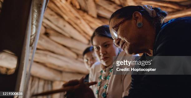 une grand-mère amérindienne (navajo) dans her sixties enseigne à ses petites-filles adolescentes comment tisser à un métier à tisser à l’intérieur dans un hogan (navajo hut) - native american ethnicity photos et images de collection
