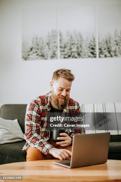 man with beard holds cup of coffee while working on laptop computer - flannel stock pictures, royalty-free photos & images