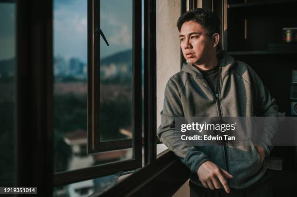 un homme chinois asiatique moyen adulte regardant à l’extérieur de la fenêtre de sa maison pendant le coucher du soleil avec l’expression faciale sérieuse avec la chemise à capuchon - homme inquiet photos et images de collection