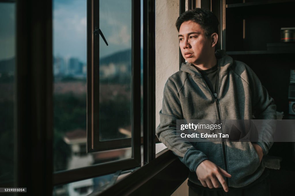 An asian chinese mid adult man looking outside of window from his home during sunset with serious facial expression with hooded shirt