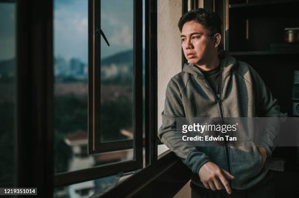 an asian chinese mid adult man looking outside of window from his home during sunset with serious facial expression with hooded shirt - depression sadness stock pictures, royalty-free photos & images