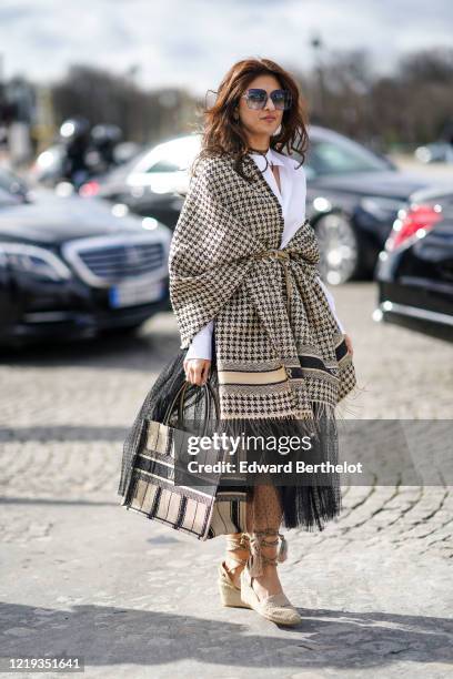 Guest wears sunglasses, a houndstooth pattern wool plaid poncho with fringes, a white shirt, a Dior shopping bag, a mesh skirt, beige shoes, outside...