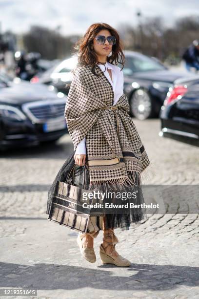 Guest wears sunglasses, a houndstooth pattern wool plaid poncho with fringes, a white shirt, a Dior shopping bag, a mesh skirt, beige shoes, outside...