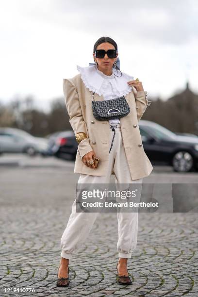 Gabriella Berdugo wears sunglasses, a white ruffled shirt, a beige oversized blazer jacket, a golden bracelet, finger rings, a Dior Saddle bag, white...