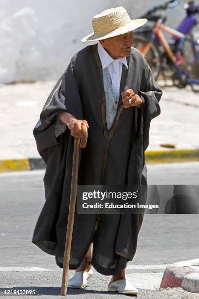 Old Berber man in traditional woolen cloak and straw hat walks with stick in street Matmata Tunisia.