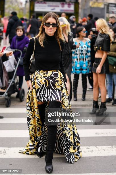 Guest wears sunglasses, a black turtleneck, a black bag with a gold chain strap, a yellow and black African design mini skirt with a full-length...