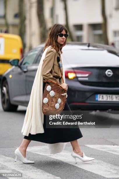 Natasha Goldenberg wears sunglasses, a cream-color knit cape, a sand-color shirt, a black skirt, white pointy heeled pumps, a Loewe bucket mesh...