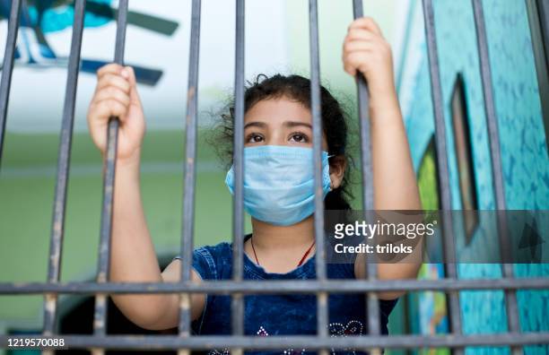 little girl wearing a face mask and peeking out from window - lockdown stock pictures, royalty-free photos & images