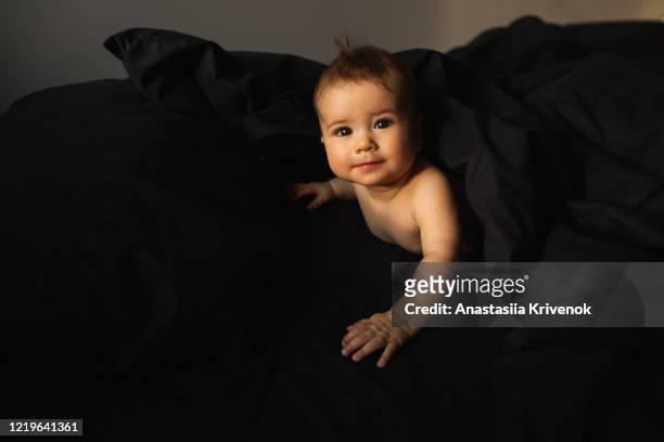 cute beautiful baby lying on black natural cotton bedding, smiling and looking at camera. portrait of crawling baby on the bed in his room. morning mood. - dark baby stock pictures, royalty-free photos & images