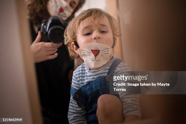 boy wearing a surgical mask painted as a cat while mom takes a photo through the mirror wearing the same mask - funny surgical masks stock pictures, royalty-free photos & images