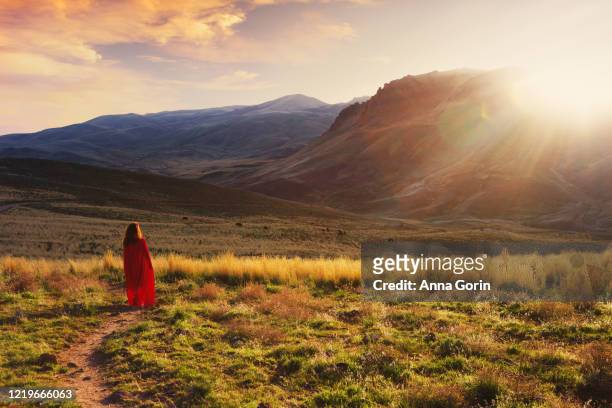back view of woman in red cape walking down path through field toward mountains at sunset - cape stock-fotos und bilder