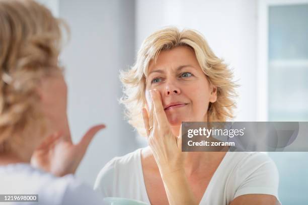 mujer adulta media extendiendo una capa de crema facial frente al espejo - mujeres de mediana edad fotografías e imágenes de stock