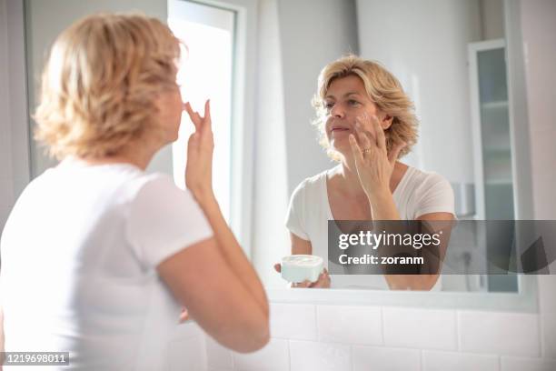 mid adult woman spreading facial cream in front of the mirror - beautiful women spreading stock-fotos und bilder
