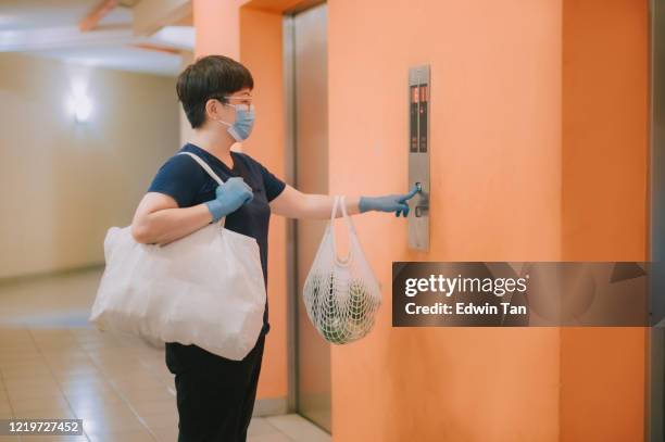 an asian chinese woman pressing the lift elevator button with her reusable bags of groceries