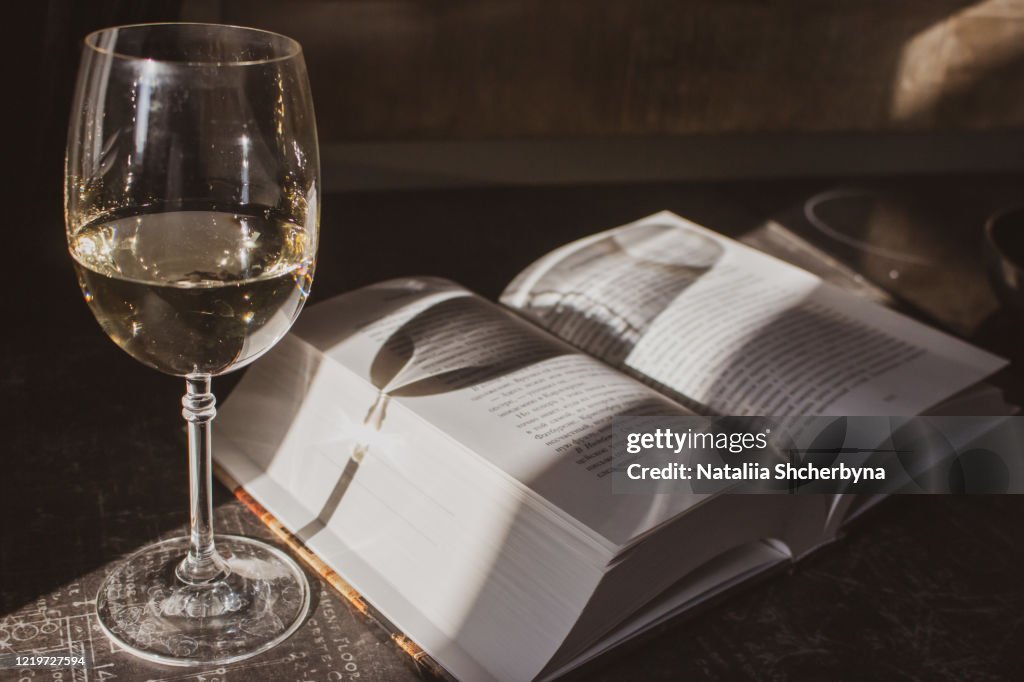 Open book and glass of white wine in sunlight filtered. Book and glass on table toned image. Literature concept. Home education concept. Read and study concept.