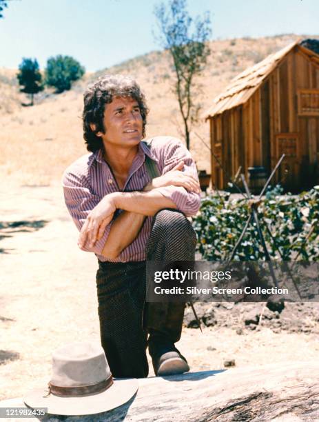 Michael Landon , US actor, poses with his arms crossed and resting on his knee with a log cabin the background in a portrait issued as publicity for...