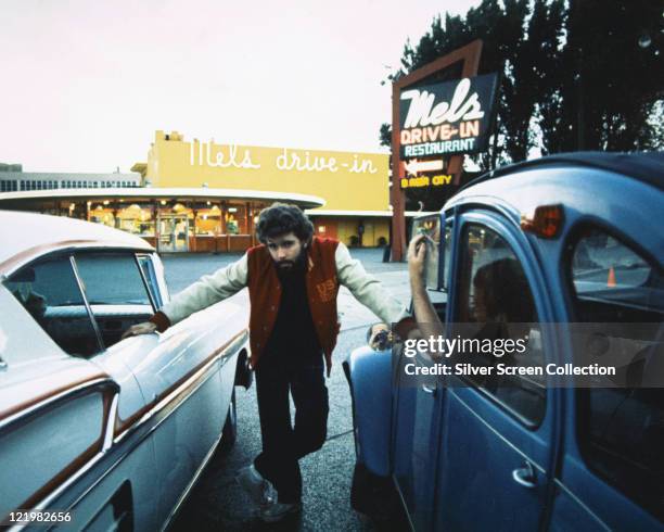 George Lucas, US film director, posing between two cars, with 'Mel's Drive-in' in the background, on the set of 'American Graffiti', San Francisco,...