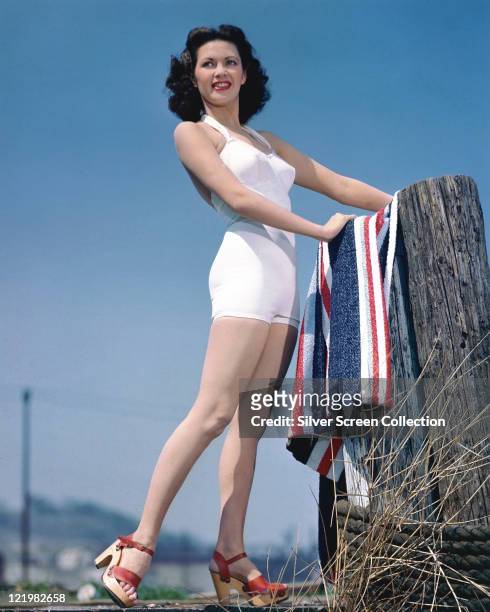 Full-length shot of Yvonne De Carlo , Canadian actress, wearing a white swimsuit while placing a red, white and blue striped towel on a wooden post,...