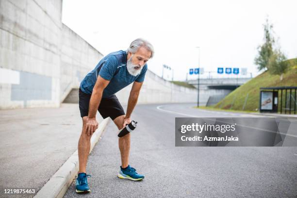 exhausted mature male runner stopping for rest and water - hand on knee stock pictures, royalty-free photos & images