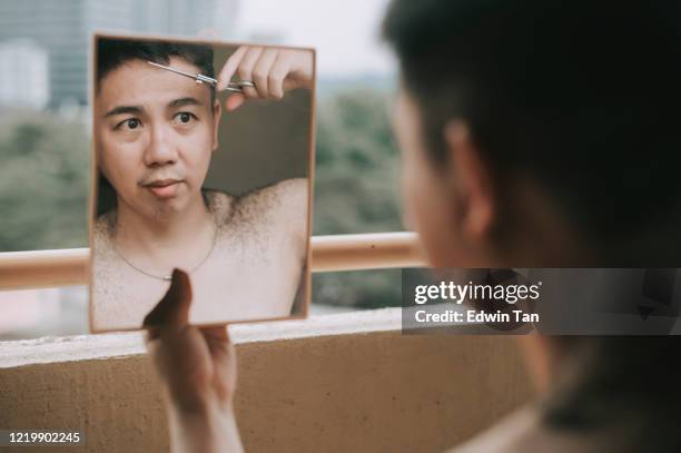 an asian chinese mid adult cutting his own hair during the quarantine at his living room balcony with a pair of scissors, mirror in the evening