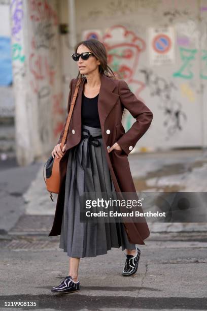 Chloe Harrouche wears sunglasses, a brown long jacket, a black top, a gray pleated skirt, a leather brown and black Loewe bag, black and white shoes,...