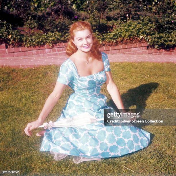 Esther Williams, US actress and swimmer, wearing a blue and white floral print dress, smiling while sitting on a patch of grass, circa 1950.