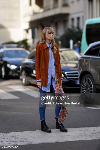 Guest wears an orange/brown suede jacket, a blue striped shirt, blue denim jeans, black shoes, a multicolor scarf with printed patterns, outside...