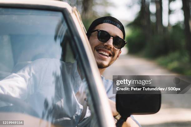 young man with black hair and sunglasses looking through window of car while driving and smiling at camera - sunny window stock pictures, royalty-free photos & images