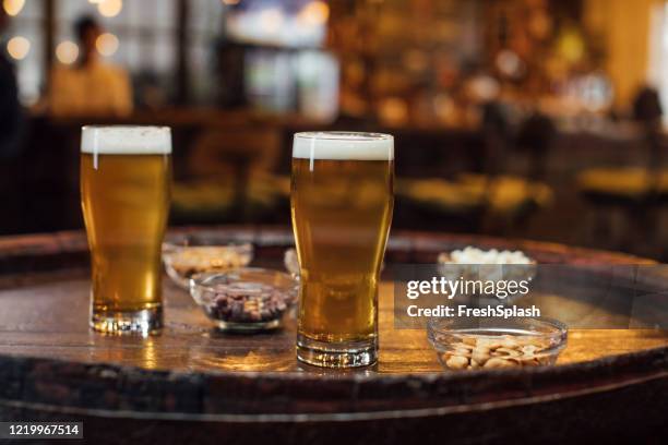 happy hour at a local pub: two glasses of light lager beer and snacks on a wooden table - lager stock pictures, royalty-free photos & images