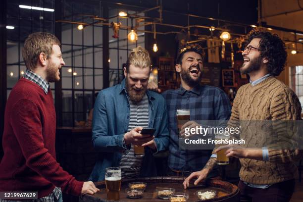 gruppe von männern, die im pub lachen, während sie bier trinken und ein fußballspiel auf dem handy sehen - bier trinken stock-fotos und bilder
