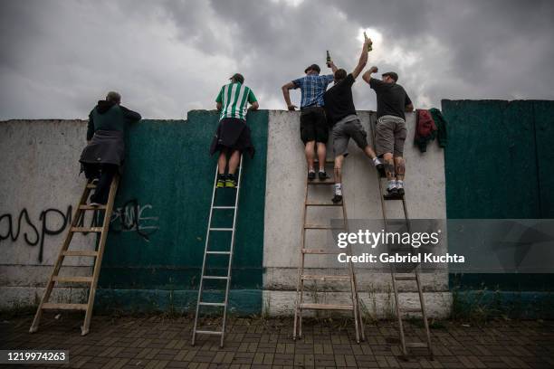 Football fans raise their drinks as they stand on ladders to peer over a wall as they watch a Czech First League match between Bohemians 1905 and FK...